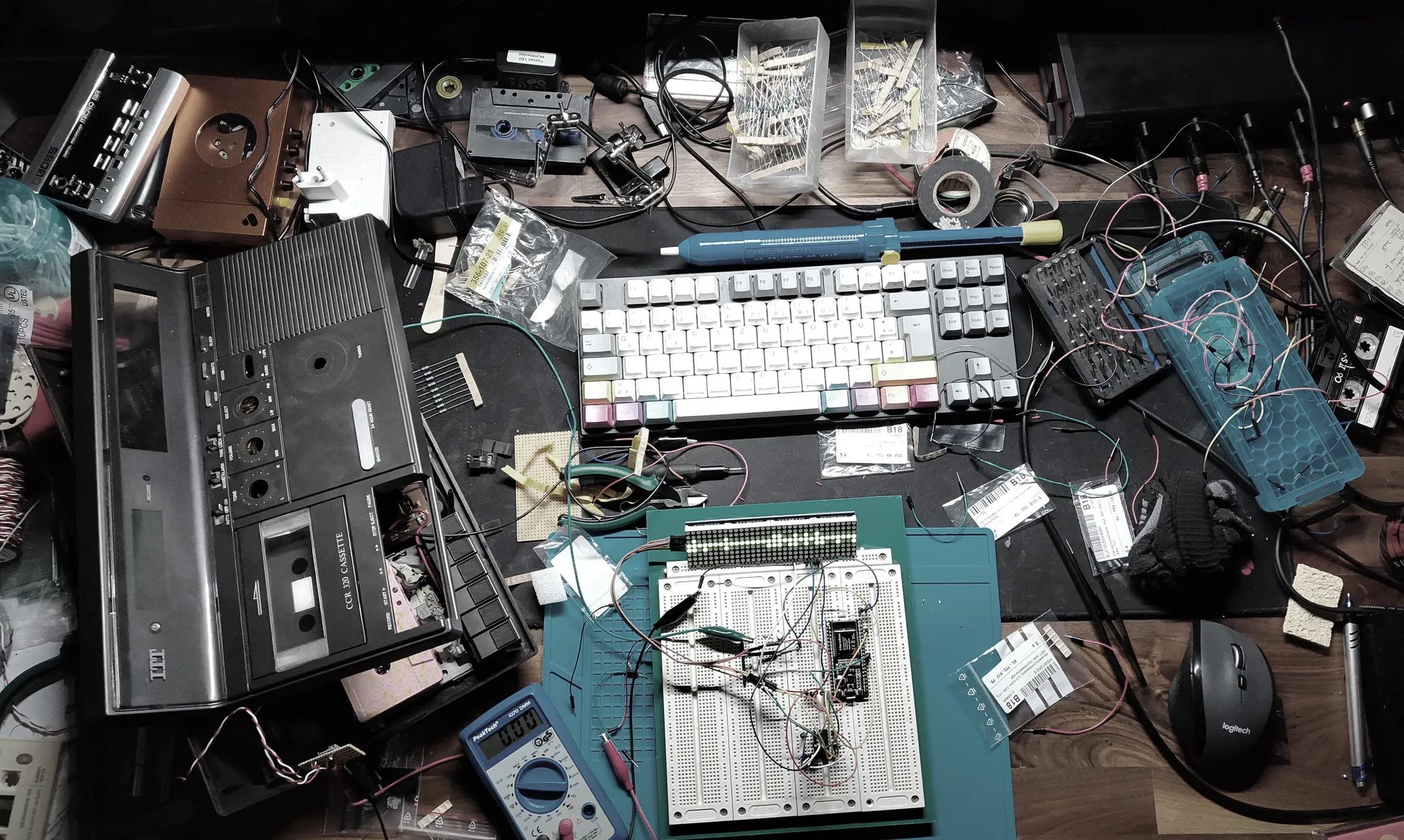 A top down photo of a very chaotic desk. There is an opened up tape cassette alarm clock, a keyboard and mouse, a multimeter, a breadboard with a wired micro-controller, some soldering tools, tape cassettes, a walkman, AC adaptors, lots of wires and some resistors. The dark wood of the desk is barely visible, because it's full of stuff.